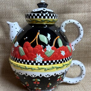 May include: A ceramic tea-for-one set featuring a teapot stacked on a teacup. The teapot has a black body with red cherries, green leaves, and white flowers, accented by a black and white checkered border and a white spout with black dots.