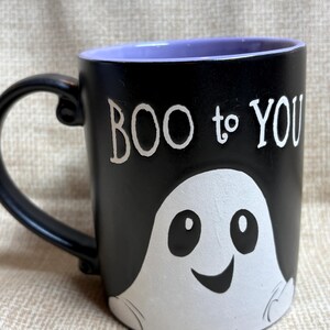 Op de afbeelding: Zwarte keramische mok met een witte spookillustratie en de tekst "BOO to YOU". De binnenkant van de mok is paars. De mok heeft een zwart handvat.