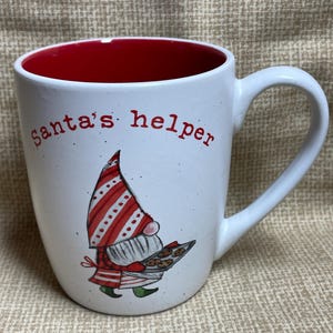 Puede incluir: Taza de cerámica blanca con interior rojo y un gnomo a rayas rojas y blancas sosteniendo una bandeja de galletas. La taza tiene el texto "Santa's helper" impreso en la parte delantera.