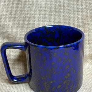 May include: A cobalt blue ceramic mug with a speckled pattern. The mug has a square handle and a glossy finish. The interior of the mug is also blue. This mug is suitable for coffee, tea, or other beverages.
