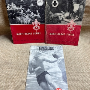 May include: Three vintage Boy Scouts of America merit badge pamphlets. The pamphlets are titled "Swimming", "First Aid", and "Lifesaving". Each pamphlet features a black and white illustration of a scout activity and the Boy Scouts of America logo.