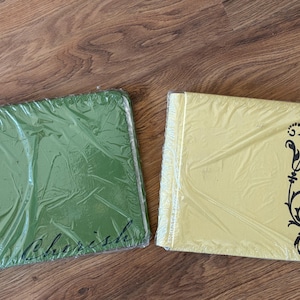 Puede incluir: Dos álbumes de fotos cuadrados, uno verde y otro amarillo, envueltos en plástico. El álbum verde tiene la palabra "Cherish" impresa. El álbum amarillo tiene un diseño floral negro en el lateral.