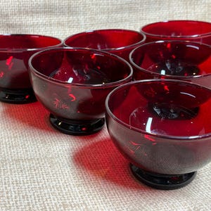 以下が含まれることがあります： 丸みを帯びた形状と黒いベースが特徴の、ルビーレッドのガラスボウル6個セット。ボウルはグループで配置され、光沢のある仕上がりとエレガントなデザインを際立たせています。デザートや少量の料理の提供に最適です。