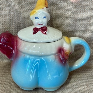 Puede incluir: Una tetera de cerámica vintage con forma de cerdo que lleva un mono azul y un sombrero amarillo. La tetera tiene una tapa extraíble con una cara caprichosa y una pajarita roja.