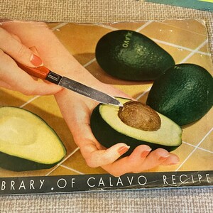 K&ouml;nnte beinhalten: Ein Vintage-Kochbuch mit gr&uuml;nem Einband mit dem Titel "Library of Calavo Recipes". Der Einband zeigt eine Nahaufnahme einer Hand, die eine Avocado mit einem Messer schneidet. Die Avocado ist gr&uuml;n und hat einen braunen Kern.