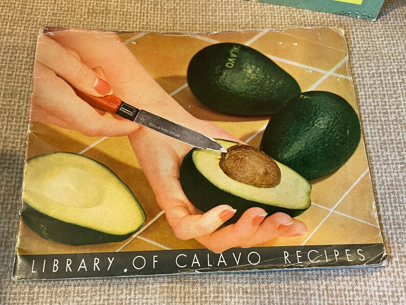 K&ouml;nnte beinhalten: Ein Vintage-Kochbuch mit gr&uuml;nem Einband mit dem Titel "Library of Calavo Recipes". Der Einband zeigt eine Nahaufnahme einer Hand, die eine Avocado mit einem Messer schneidet. Die Avocado ist gr&uuml;n und hat einen braunen Kern.