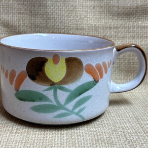 May include: A ceramic soup mug with a floral design. The mug is white with brown speckles and a brown handle. The floral design features brown, orange, and green colors. The mug is approximately 4 inches in diameter and 3 inches tall.