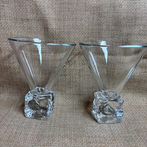 May include: Two clear glass martini glasses. The glasses have a triangular bowl shape and sit atop a square, textured base. The background is a neutral tan fabric.