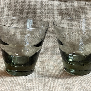 May include: Two clear glass tumblers with a dark green tint. The glasses have a rounded, slightly flared shape.