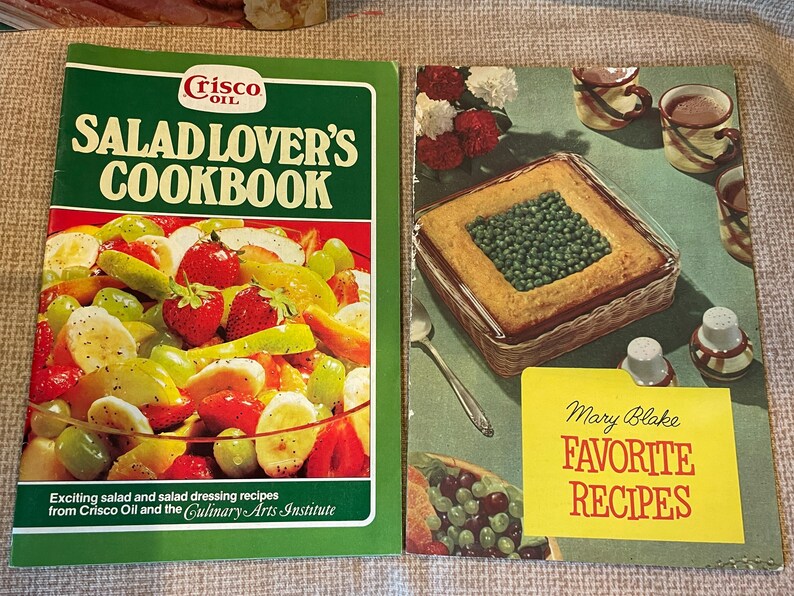 K&ouml;nnte beinhalten: Zwei Vintage-Kochb&uuml;cher, eines mit dem Titel "Salad Lover's Cookbook" mit einem Foto eines Obstsalats auf dem Cover und das andere mit dem Titel "Mary Blake Favorite Recipes" mit einem Foto einer Auflaufform gef&uuml;llt mit Erbsen auf dem Cover.