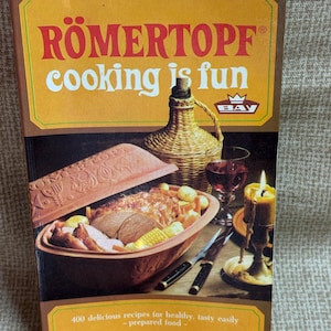 May include: A vintage cookbook titled "RÖMERTOPF cooking is fun" with a brown cover. The cover features a terracotta cooking pot filled with food, a wicker-covered bottle, a lit candle, and a glass of red wine. The book promises 400 recipes.