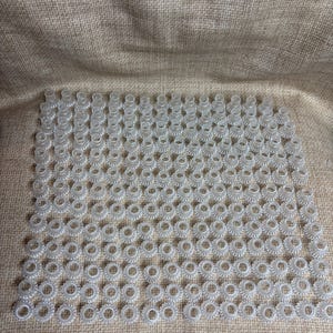 May include: A large collection of small, clear plastic gears arranged in a grid pattern. Each gear has a circular shape with a toothed outer edge and a central hole. The gears are displayed on a textured, tan fabric surface.