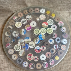 Puede incluir: Un estuche de costura circular transparente lleno de numerosos carretes de hilo de varios colores y tamaños. Los carretes están dispuestos en compartimentos individuales, mostrando una variedad de marcas y diseños de hilo. Ideal para coser y manualidades.