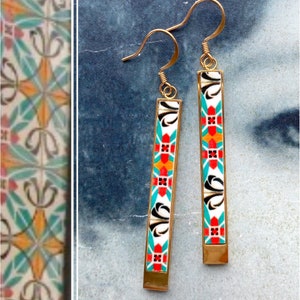 May include: A pair of gold dangle earrings with a white, black, red, orange, and turquoise geometric pattern.