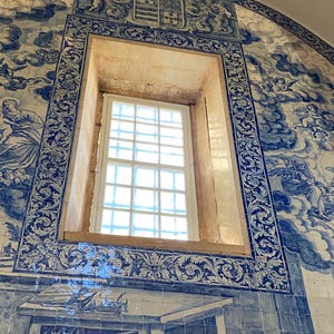 May include: A rectangular window set within a wall covered in blue and white azulejo tiles. The tiles feature intricate illustrations of clouds, figures, and a central coat of arms above the window frame, with a grid-patterned window pane.