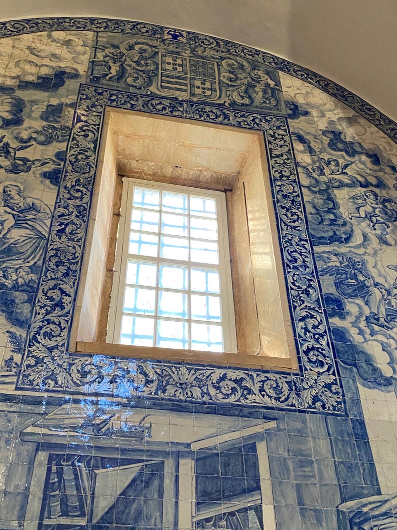 May include: A rectangular window set within a wall covered in blue and white azulejo tiles. The tiles feature intricate illustrations of clouds, figures, and a central coat of arms above the window frame, with a grid-patterned window pane.