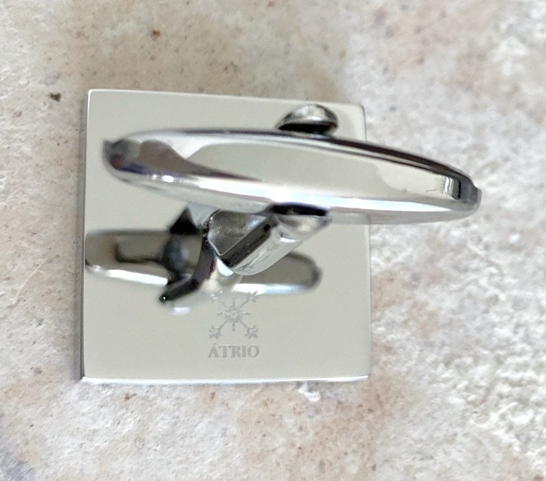 May include: Silver cufflinks with a square base and a rounded top. The cufflinks have a logo engraved on the base that says "ATRIO" with a starburst design.