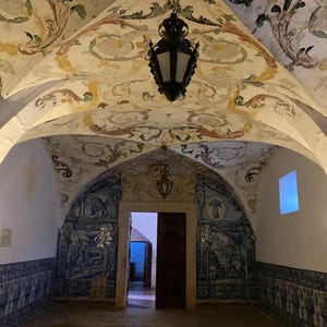 May include: An interior room featuring a vaulted ceiling with painted floral motifs in muted tones. The lower walls are covered in blue and white azulejo tiles depicting historical scenes. A dark metal lantern hangs from the center of the ceiling.