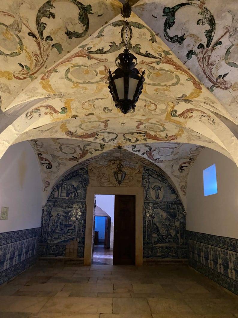 May include: An interior room featuring a vaulted ceiling with painted floral motifs in muted tones. The lower walls are covered in blue and white azulejo tiles depicting historical scenes. A dark metal lantern hangs from the center of the ceiling.