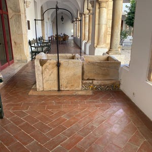May include: A rectangular stone basin with a central metal arch structure, positioned on a terracotta tiled floor within a covered walkway featuring stone columns and arched ceilings. The base of the basin is decorated with patterned tiles.