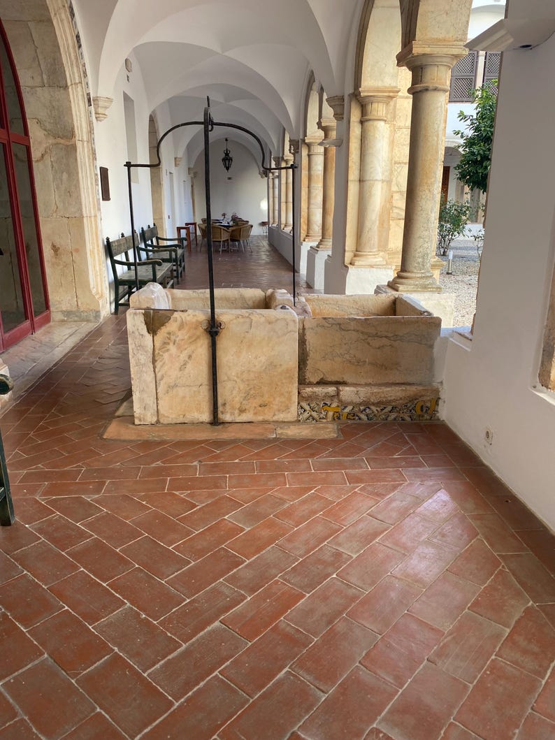 May include: A rectangular stone basin with a central metal arch structure, positioned on a terracotta tiled floor within a covered walkway featuring stone columns and arched ceilings. The base of the basin is decorated with patterned tiles.