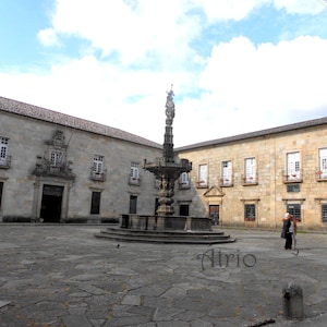 May include: A stone fountain with a statue on top, situated in a courtyard with a stone floor and buildings on all sides.
