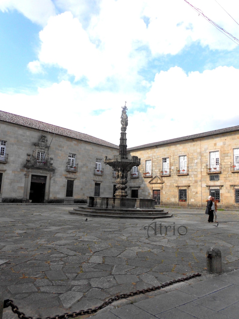 May include: A stone fountain with a statue on top, situated in a courtyard with a stone floor and buildings on all sides.
