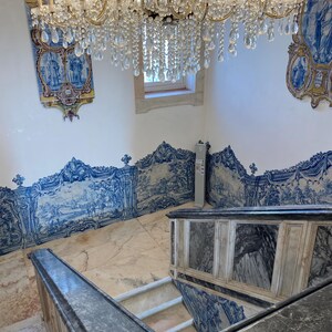 May include: A grand staircase featuring dark grey marble railings and steps, surrounded by walls adorned with intricate blue and white azulejo tiles depicting landscape scenes. A large, multi-tiered crystal chandelier hangs above the stairwell.