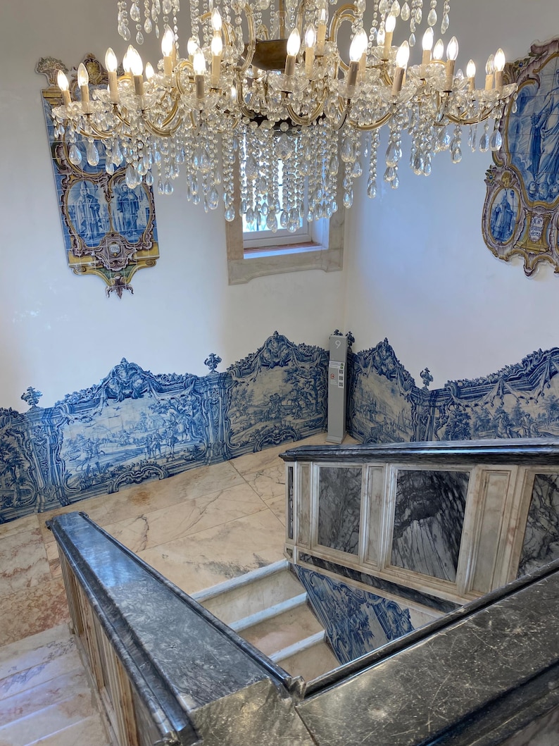May include: A grand staircase featuring dark grey marble railings and steps, surrounded by walls adorned with intricate blue and white azulejo tiles depicting landscape scenes. A large, multi-tiered crystal chandelier hangs above the stairwell.