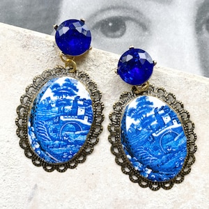 May include: A pair of vintage-style earrings. Each earring features a blue gemstone stud, connected to an oval pendant with a blue and white landscape design. The pendants are framed by a decorative, bronze-colored filigree.