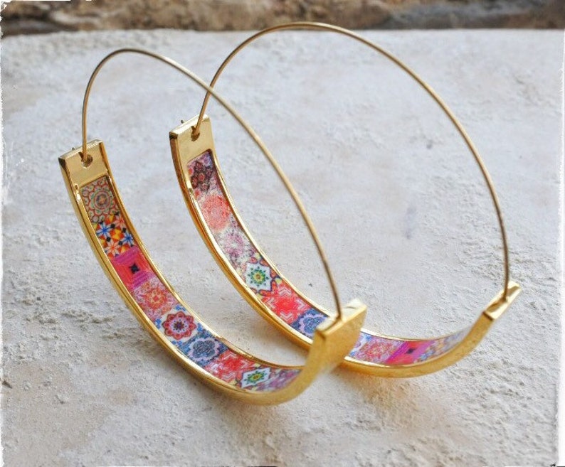 May include: A pair of gold hoop earrings with a colorful patterned design. The pattern features a variety of geometric shapes and colors, including pink, blue, and green.