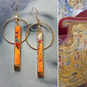 May include: A pair of gold hoop earrings with a long, rectangular dangle. The dangles are orange with a floral pattern.