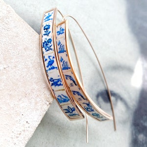 Op de afbeelding: Een paar gouden creolen met een wit en blauw patroon. Het patroon bevat vogels, bloemen en geometrische vormen.