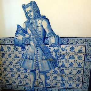 Op de afbeelding: Een blauwe en witte tegelmuur met daarop een man in een blauw-witte gepatroneerde jas en broek. De man houdt een staf vast en staat voor een betegelde muur met een bloemenontwerp.