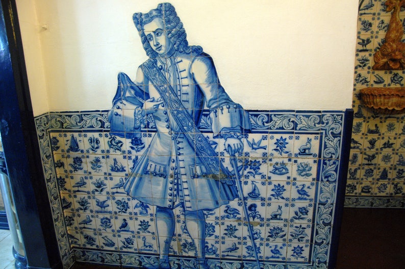 Op de afbeelding: Een blauwe en witte tegelmuur met daarop een man in een blauw-witte gepatroneerde jas en broek. De man houdt een staf vast en staat voor een betegelde muur met een bloemenontwerp.