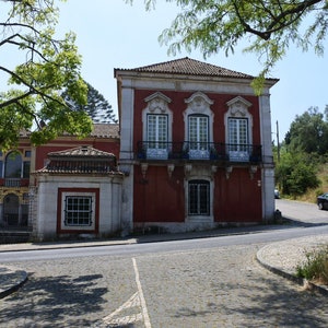 May include: A red brick building with white trim and a tiled roof. The building has a balcony with black railings and windows with white frames. The building is located on a street with a cobblestone road.