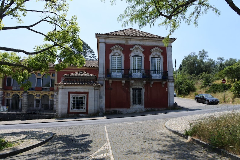 May include: A red brick building with white trim and a tiled roof. The building has a balcony with black railings and windows with white frames. The building is located on a street with a cobblestone road.