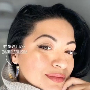 May include: A woman with dark hair wearing a gray turtleneck sweater and gold hoop earrings with blue and white accents. The text "MY NEW LOVES @ATRIOAZULEJOS" is visible in the image.