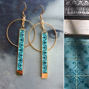 May include: Gold hoop earrings with a turquoise and black patterned bar pendant. The pattern is a repeating floral design.