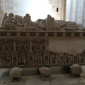 May include: A detailed stone tomb with intricate carvings depicting figures and scenes. The tomb is resting on four lion-like feet and features a raised section with a series of arches.