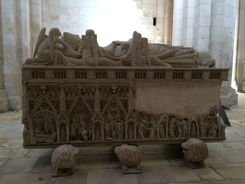May include: A detailed stone tomb with intricate carvings depicting figures and scenes. The tomb is resting on four lion-like feet and features a raised section with a series of arches.