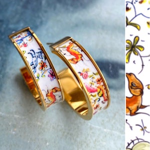 Könnte beinhalten: Ein Paar goldene Creolen mit weißem Hintergrund und bunten floralen und tierischen Designs.