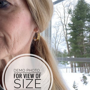 May include: A close-up of a person's ear wearing a gold hoop earring. The earring is a simple, round hoop with a smooth finish. The background is blurred, showing a window with a view of a snowy landscape.