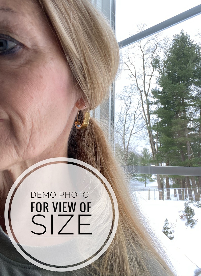 May include: A close-up of a person's ear wearing a gold hoop earring. The earring is a simple, round hoop with a smooth finish. The background is blurred, showing a window with a view of a snowy landscape.