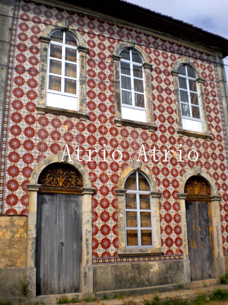 May include: A building with a red and white tiled facade. The building has three windows with white frames and a wooden door with a wrought iron gate. The text "Atrio Atrio" is visible on the building.