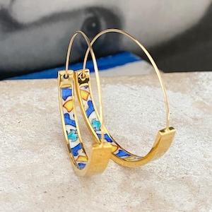 May include: Gold-tone hoop earrings with a colorful, patterned design. The earrings feature a vibrant mix of blue, yellow, and white geometric shapes. The hoops have a sleek, modern design, perfect for adding a pop of color to any outfit.