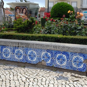 May include: A stone bench with blue and white tile accents sits in front of a lush green hedge. The bench is made of gray stone and has a smooth, polished surface. The tiles are arranged in a repeating pattern and feature a floral design.