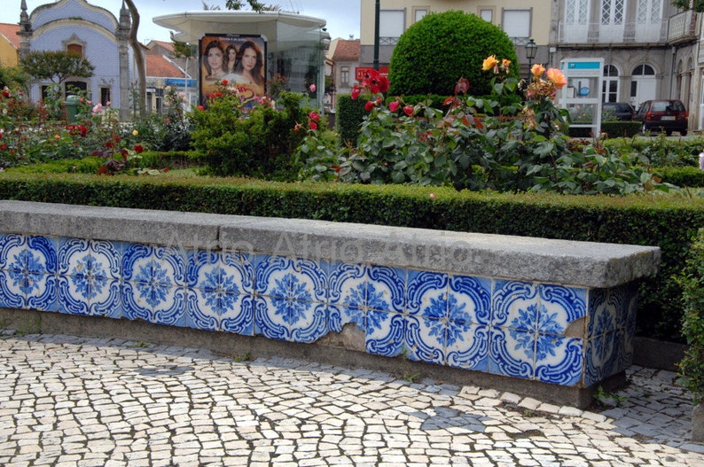 May include: A stone bench with blue and white tile accents sits in front of a lush green hedge. The bench is made of gray stone and has a smooth, polished surface. The tiles are arranged in a repeating pattern and feature a floral design.