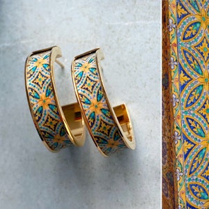 Könnte beinhalten: Ein Paar goldene Creolen mit einem blauen und gelben Muster. Das Muster erinnert an traditionelle Fliesenarbeiten.