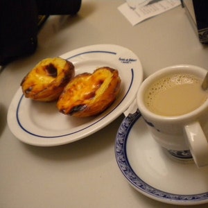 Puede incluir: Dos tartas de crema portuguesas doradas sobre un plato blanco con borde azul. Una taza de caf&eacute; con leche en una taza blanca con borde azul se encuentra sobre un platillo blanco con un patr&oacute;n azul y blanco.